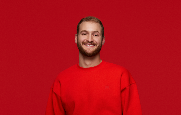 Ein junger Mann in einem roten Pullover blickt lächelnd in die Kamera.
