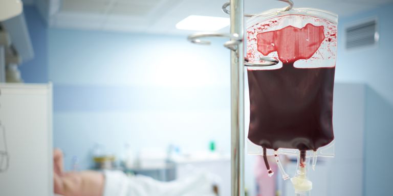 Chambre d'hôpital avec une poche de sang, à l'arrière-plan la silhouette floue d'un patient.