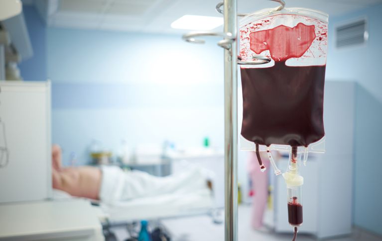 Chambre d'hôpital avec une poche de sang, à l'arrière-plan la silhouette floue d'un patient.